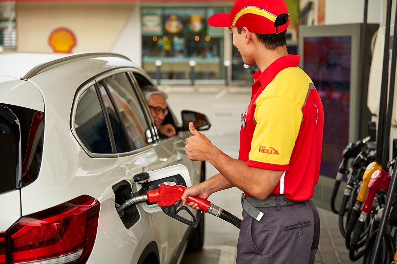 Empleado de Shell con uniforme rojo y amarillo llena el tanque de un vehículo blanco mientras conversa con un cliente en una estación de servicio. Servicio al cliente y abastecimiento de combustible en estaciones Shell.